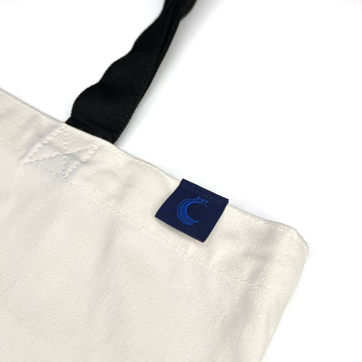 Close-up of the navy woven Sapphire Shadow Studio label stitched into the tote's top hem, with the canvas weave, the hem topstitching and one black strap in frame.