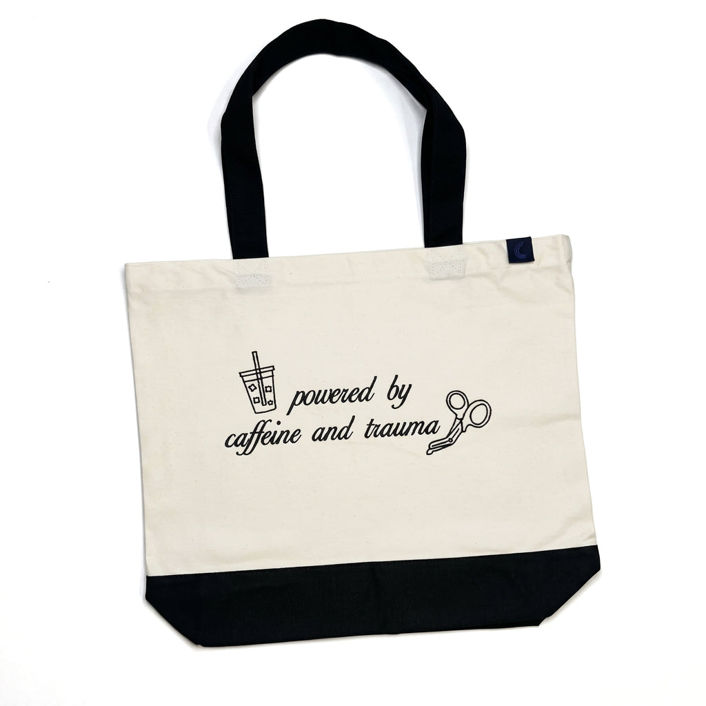 Natural canvas tote bag with a black base and black shoulder straps, laid flat, embroidered on the front with an iced drink, a pair of trauma shears, and the words powered by caffeine and trauma.