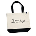 Natural canvas tote bag with a black base and black shoulder straps, laid flat, embroidered on the front with an iced drink, a pair of trauma shears, and the words powered by caffeine and trauma.