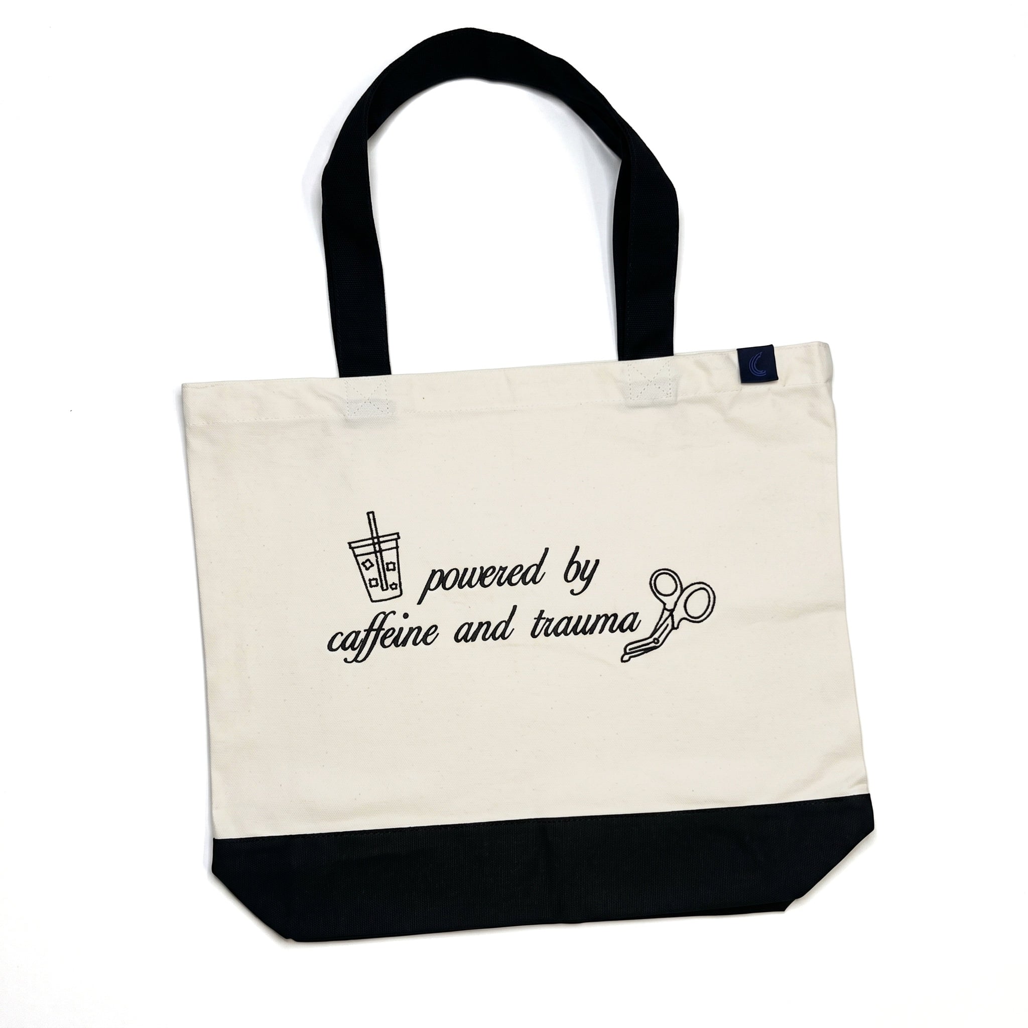 Natural canvas tote bag with a black base and black shoulder straps, laid flat, embroidered on the front with an iced drink, a pair of trauma shears, and the words powered by caffeine and trauma.
