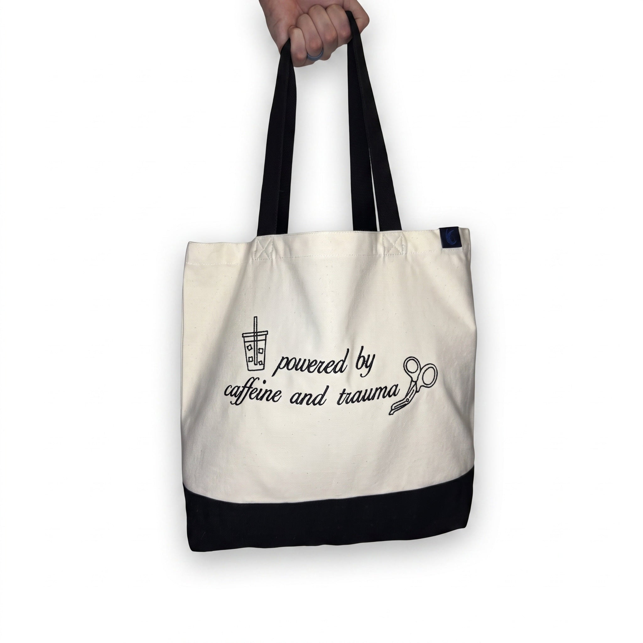 The natural canvas tote held up by its two black straps, showing how it hangs and how deep it sits when carried, with the Caffeine and Trauma embroidery across the front.
