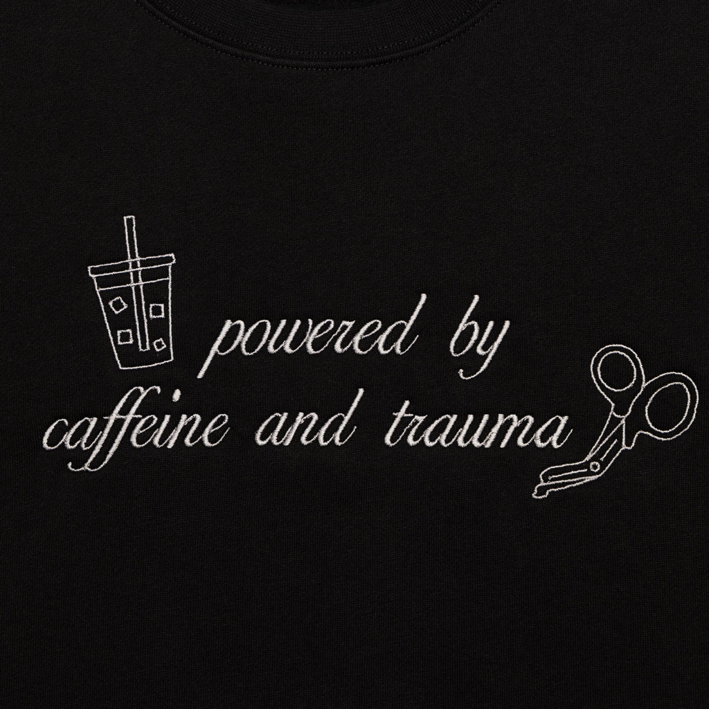 Close-up of the Powered by Caffeine and Trauma design embroidered on the chest of the Black crewneck
