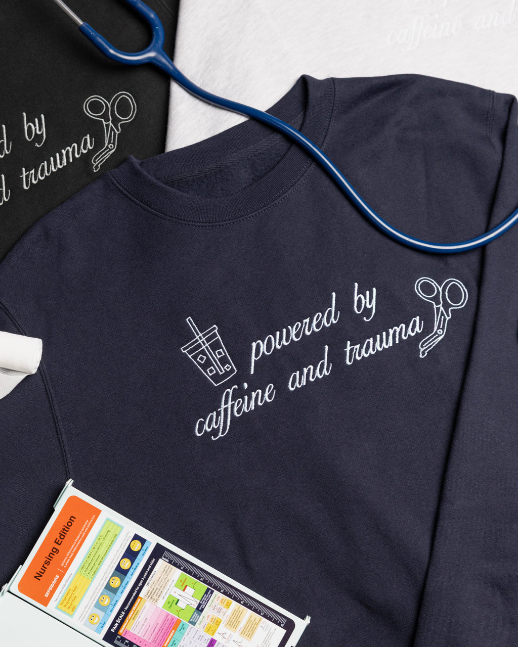 Shift Fuel crewneck styled with a stethoscope, bandage roll, and nursing reference card, with additional crewnecks in the background