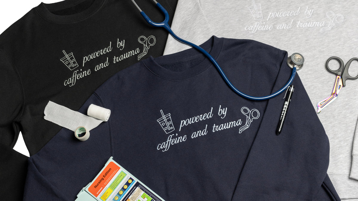 Three folded Shift Fuel crewnecks styled together with a stethoscope, medical tape, a pen, trauma shears, and a nursing reference card